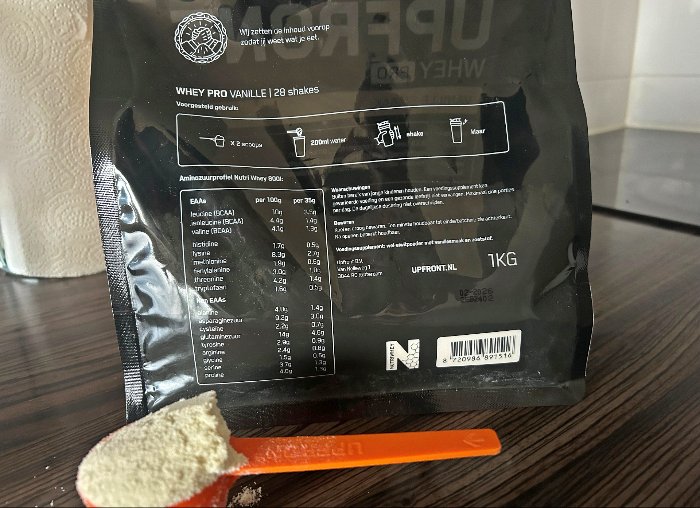 De achterkant van een zak Upfront Whey PRO met voor de zak een met whey gevulde oranje Upfront scoop