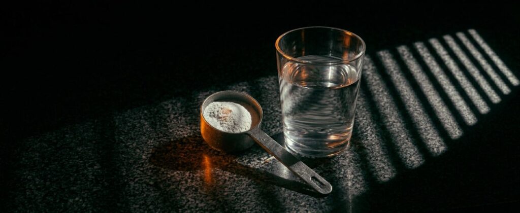 Creatine monohydraat in een maatschep naast een glas water op een donker aanrechtblad, klaar voor dagelijks gebruik